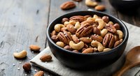 Mixed nuts in dark bowl on rustic wooden table with scattered cashews, pecans, almonds and walnuts for healthy snacking and cooking ingredients.