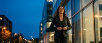 Confident businesswoman in black suit walking along modern office building at dusk in urban financial district with city lights and glass architecture.