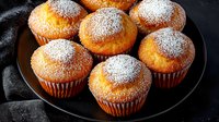 Fresh homemade vanilla muffins dusted with powdered sugar served on black plate against dark background, close up view of sweet pastry dessert.