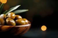 Fresh green olives in wooden bowl on dark background with bokeh lights and olive leaves, moody food photography composition for Mediterranean cuisine.