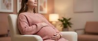 Pregnant woman in pink knit dress relaxing in modern living room chair, expecting mother maternity lifestyle concept for healthcare and family planning.