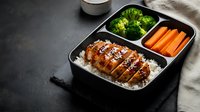 Japanese bento lunch box with teriyaki chicken over rice, steamed broccoli and carrot sticks in compartments on dark surface for meal prep and healthy eating.