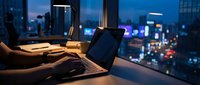 Professional working late at night on laptop in modern office with city lights view through window creating productive evening work atmosphere.