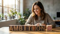 Woman with long brown hair, Caucasian ethnicity, smiling while sitting at desk with wooden blocks spelling ambition and target symbol, modern office with plants and natural light.
