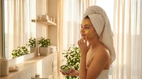 Young African American woman applying skincare routine in bright bathroom with towel wrapped around head after shower for wellness and self-care concepts.
