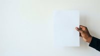 Person with medium skin tone holding blank white paper against neutral wall background for mockup and branding purposes.