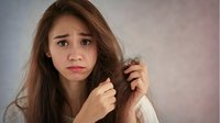 Young Asian woman with concerned expression examining damaged, dry hair ends against soft pink background, demonstrating hair care problems and split ends.