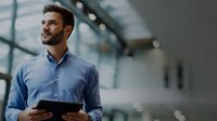 Young businessman with beard holding digital tablet in modern office building, looking thoughtfully at distance with confident expression.