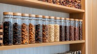 Glass storage jars filled with various nuts, dried fruits and coffee beans arranged neatly on wooden pantry shelves, showcasing modern kitchen organization and bulk food storage.