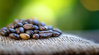 Fresh roasted coffee beans scattered on natural burlap fabric with soft green bokeh background, macro photography showing rich texture and detail.