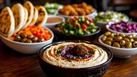 Creamy hummus bowl garnished with herbs and spices, surrounded by Mediterranean mezze platter including pita bread, olives, fresh vegetables and appetizers on wooden table.