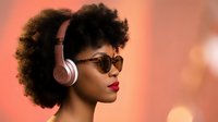 Young African American woman with curly afro hair wearing sunglasses and wireless headphones against warm bokeh background for music and lifestyle content.