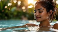 Young Hispanic woman relaxing in swimming pool with eyes closed during sunset, water droplets on face, natural bokeh background creates dreamy atmosphere.
