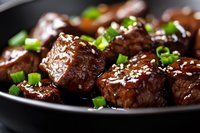 Glazed beef chunks with sesame seeds and green onions served on black plate, close up view of Asian style meat dish with sweet and savory sauce.