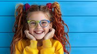 Cheerful young girl with red pigtails and colorful glasses wearing yellow sweater, expressing joy and happiness against bright blue wooden background.