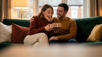 Happy young Caucasian couple sharing gift box on cozy living room sofa during romantic evening at home together.