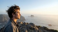 Young man with curly hair gazing at ocean sunset from rocky cliff, contemplating future with peaceful expression, adventure and freedom concept.