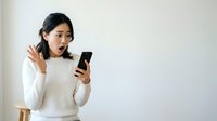 Young Asian woman with dark hair wearing white sweater, holding smartphone with surprised expression, isolated on white background for advertising and digital communication concepts.
