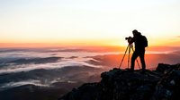 Male photographer silhouette capturing sunrise from mountain peak above clouds with professional camera and tripod equipment for landscape photography adventure.
