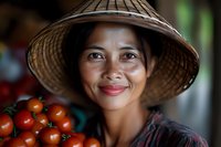 Smiling Asian woman vendor wearing traditional conical hat holds fresh red tomatoes at local market, authentic cultural portrait showing genuine warmth and hospitality.