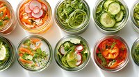Fresh pickled vegetables in glass jars viewed from above. Colorful assortment includes radishes, carrots, cucumbers and herbs in brine for fermentation.