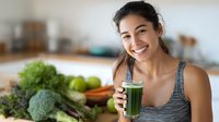 Happy young Asian woman drinking fresh green vegetable juice smoothie in modern kitchen with healthy organic produce on counter for wellness lifestyle.