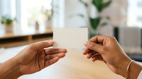 Two hands exchanging blank white card in bright office environment with plants and windows, suitable for business, corporate identity, networking, and professional communication concepts.