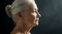 Senior woman with gray hair in bun, profile view, mature facial features, age spots, wrinkles, contemplative expression against dark background for healthcare and aging concepts.