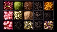 Colorful collection of dried flowers, tea leaves, spices and herbs in black square compartments arranged in grid pattern, overhead view on dark background.