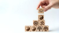 Hand placing wooden blocks with spiritual symbols including yin yang, lotus, om, and tree of life in pyramid formation on white background.