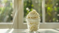 Creamy vanilla soft serve ice cream swirled in clear plastic cup against sunlit window with bokeh background, fresh dairy dessert with smooth texture.