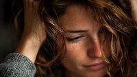 Young blonde woman with freckles holding head in hands showing stress, anxiety or headache in warm natural lighting for mental health concepts.