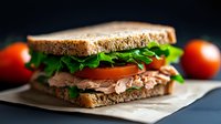 Fresh tuna sandwich with tomato slices, crisp arugula on whole grain bread, served on parchment paper against dark background with fresh tomatoes.