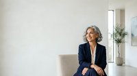 Senior woman with gray hair wearing navy blazer and white blouse, sitting in modern office chair against white wall background, suitable for corporate, business, and professional website design.