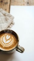 Steaming hot latte with heart-shaped foam art in ceramic mug on white surface with cozy knit sweater and rustic wooden background for cafe branding.