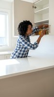 Young African American woman organizing kitchen pantry shelves in modern white cabinet wearing blue plaid shirt during home renovation project.