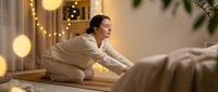 Young Asian woman practicing yoga meditation in cozy bedroom with warm lighting and string lights creating peaceful wellness atmosphere for relaxation.
