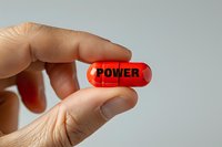 Red transparent capsule pill with power text held between fingers against neutral background, representing strength, enhancement or medication concept.