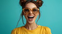 Young woman with space buns hairstyle wearing yellow t-shirt and oversized sunglasses expressing extreme joy and excitement against turquoise background.