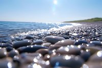 Smooth pebbles on beach shore with sunlight sparkling on wet stones and ocean waves, peaceful seascape view at sunny day with blue water and sky.