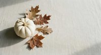 White pumpkin with dried autumn oak leaves on neutral linen fabric background for seasonal home decor and fall harvest celebrations.