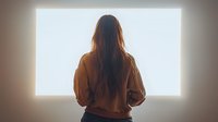 Young woman with long dark hair wearing brown sweater standing in front of bright glowing window, view from behind creating atmospheric silhouette effect.