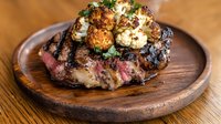 Juicy grilled ribeye steak topped with roasted cauliflower florets and fresh herbs, served medium rare on rustic wooden board. Perfect for restaurant menu.
