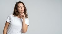 Thoughtful young Caucasian woman in white t-shirt touching chin while looking up, contemplating decision on neutral background for marketing campaigns.