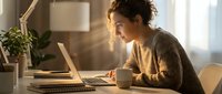 Young woman with curly hair working on laptop at home office desk with coffee cup and plant in warm natural lighting for remote work productivity.