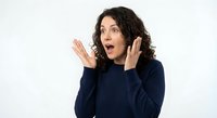Surprised young Hispanic woman with curly hair raising hands in shock gesture wearing navy blue sweater on white background for marketing design.