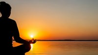 Silhouette of woman meditating in lotus pose at sunset by calm lake with golden sky reflection on water creating peaceful zen atmosphere for wellness.