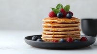 Stack of golden pancakes topped with fresh raspberries and blueberries, drizzled with maple syrup on dark plate against light background, morning breakfast setup.