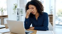 Stressed young African American businesswoman with headache sitting at office desk with laptop experiencing work burnout and fatigue.