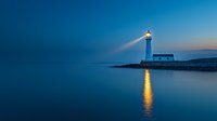 Lighthouse beam reflecting on calm ocean water at dusk, illuminated white tower against deep blue twilight sky creates serene coastal scene.